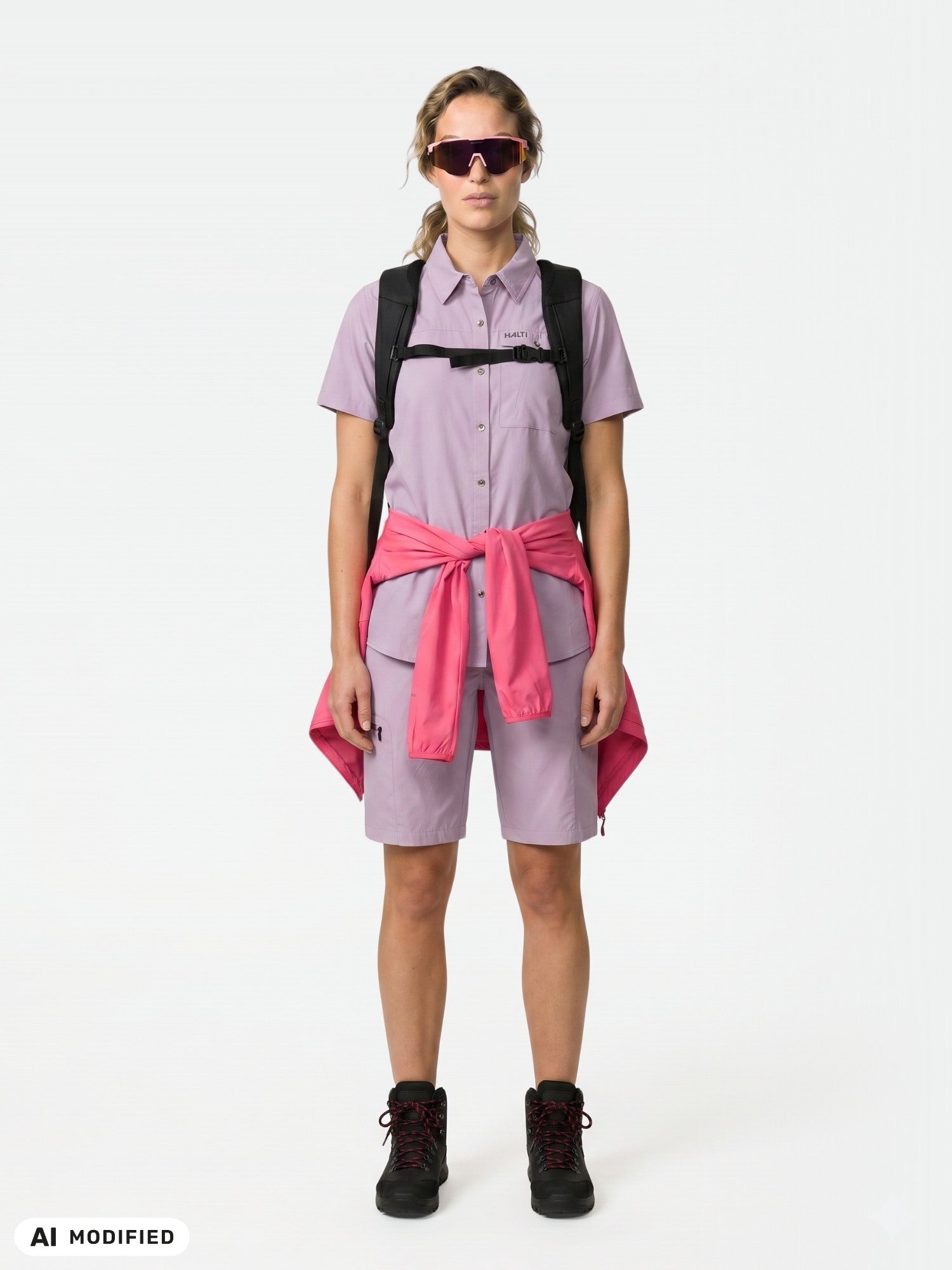 Woman in Halti Pallas Cool Shorts, pink jacket at waist, sunglasses, backpack, black boots, facing forward.