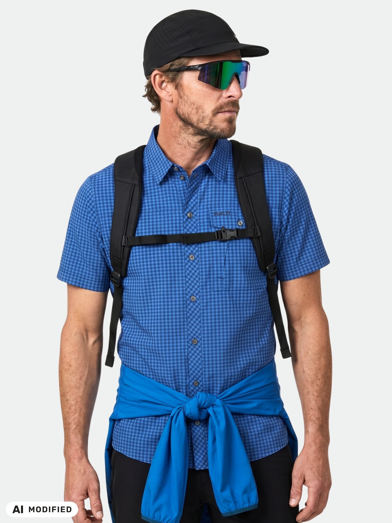 Man in Halti Samoy Trekking Shirt (Men's), sunglasses, black cap, backpack, blue jacket tied at waist.