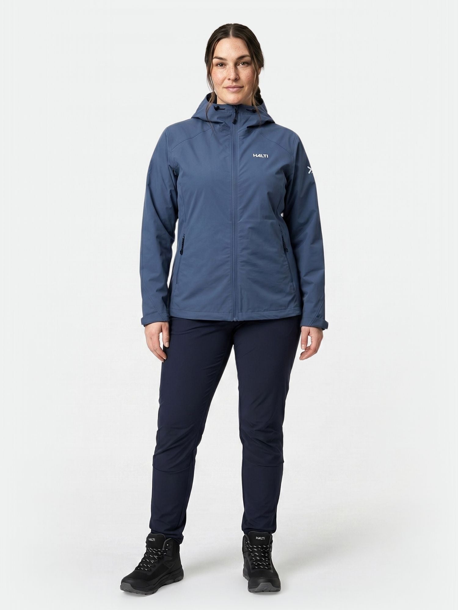 A person faces forward in a Halti Finholma X-stretch Women's Plus outdoor jacket, blue hood, dark pants, and black boots.