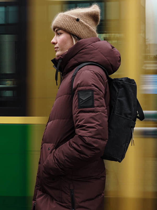 Woman in Halti Linjat Parka Jacket and tan beanie stands in profile with black backpack, blurred yellow-green background.