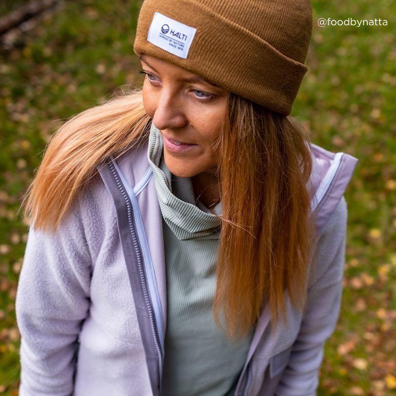 A woman wearing the Halti Laus Beanie and a light jacket stands on grass outdoors, looking to the side.