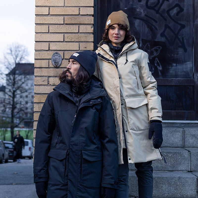 Two people in Halti Bergga Womens DrymaxX Winter Parkas and winter hats stand by a graffiti-covered brick wall.