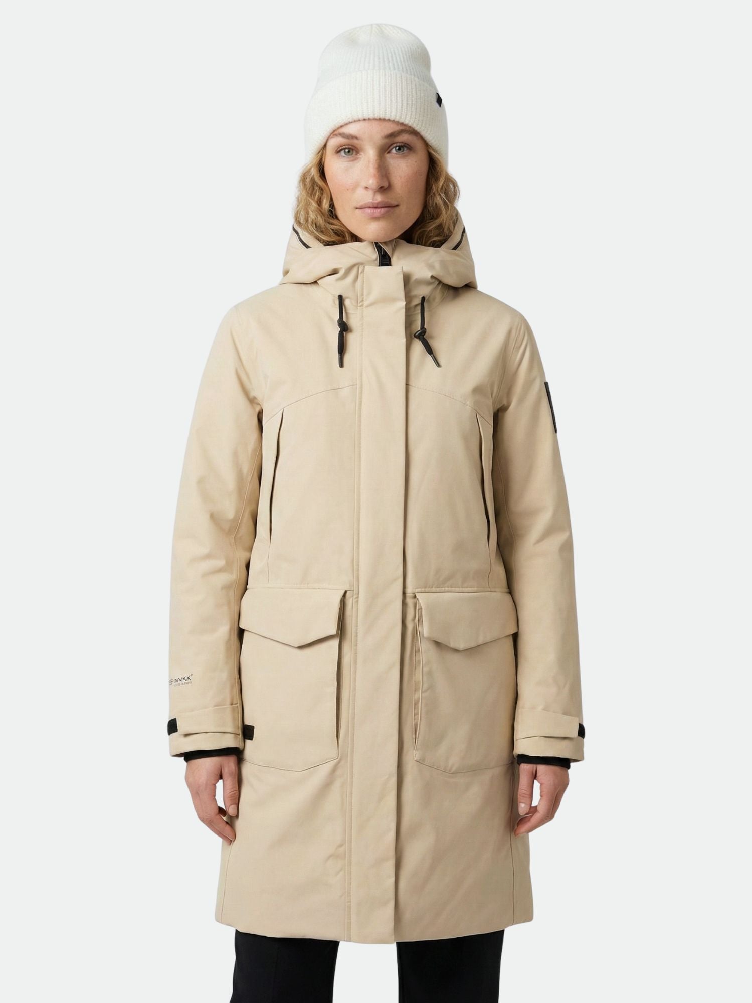 A person wearing the Halti Bergga Women's DrymaxX Winter Parka in beige and a white knit beanie, on a light background.
