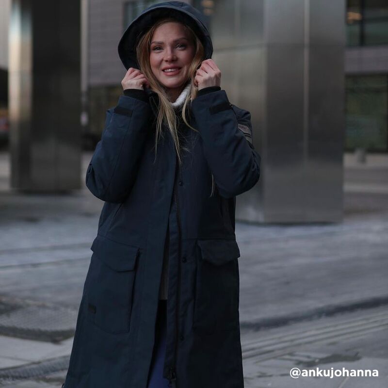 A woman in a Halti Bergga Womens DrymaxX Winter Parka stands outdoors, hood up, with city buildings behind her.