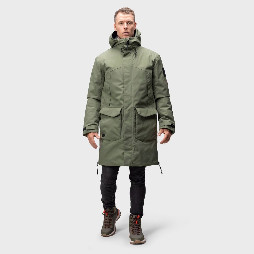 A man wears the Halti Bergga Mens DrymaxX Winter Jacket in green, paired with black pants and hiking boots.