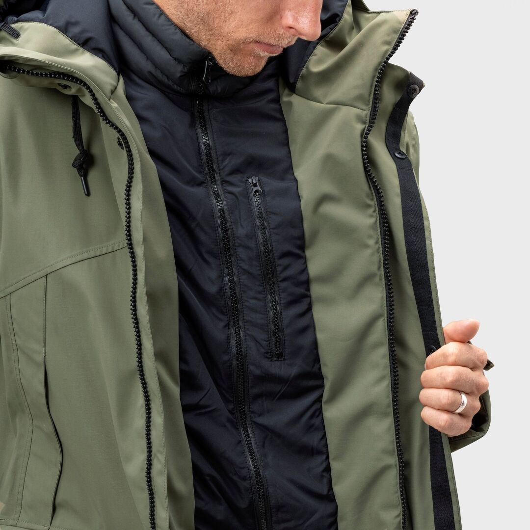 A person wears a green Halti Bergga DX Winter Parka Men's over a black jacket, holding it open to show the inner layer.