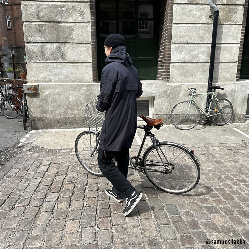 Someone wearing the Halti Kurvi Mens DrymaxX 3L Parka Jacket walks a bike along a cobblestone street by a brick building.