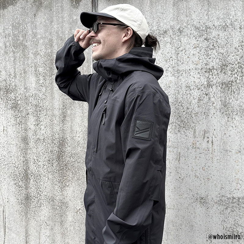 A person in the Halti Kurvi Mens DrymaxX 3L Parka Jacket, a light cap, and sunglasses smiles by a concrete wall.