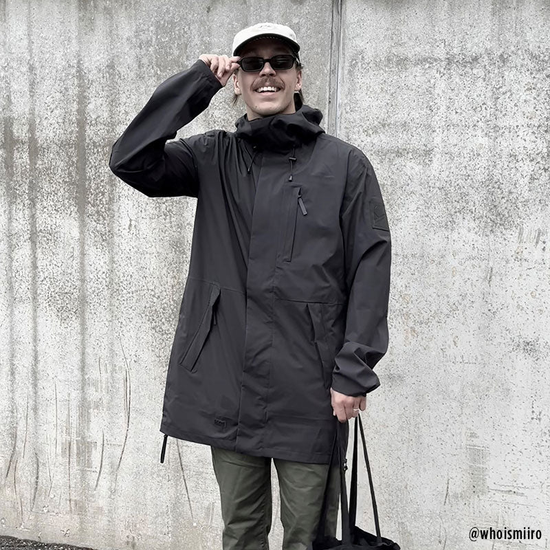 A person in a Halti Kurvi Mens DrymaxX 3L Parka stands by a concrete wall, wearing sunglasses and holding a black bag.