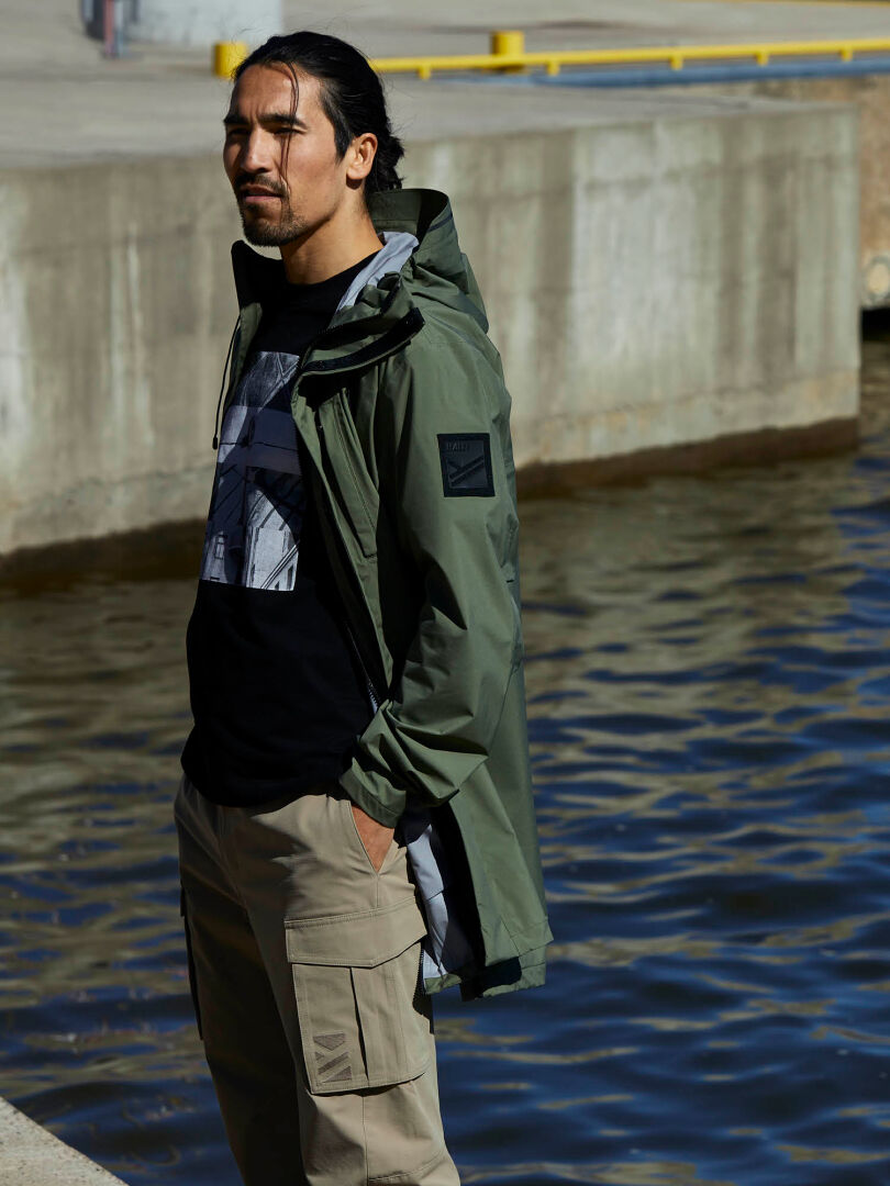 A person in a Halti Kurvi Mens DrymaxX 3L Parka Jacket with reflective details stands by water on a sunny day.