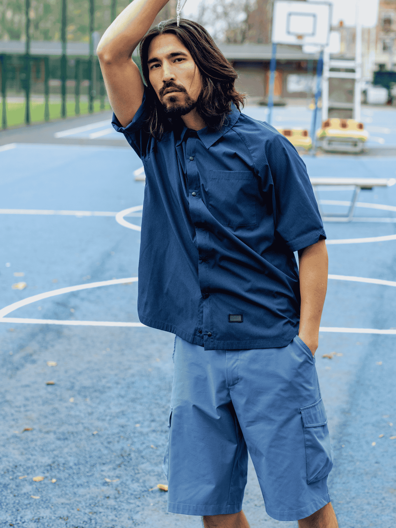 Man with long hair wearing the Halti Baana Unisex Shirt stands on an outdoor basketball court with one arm raised.