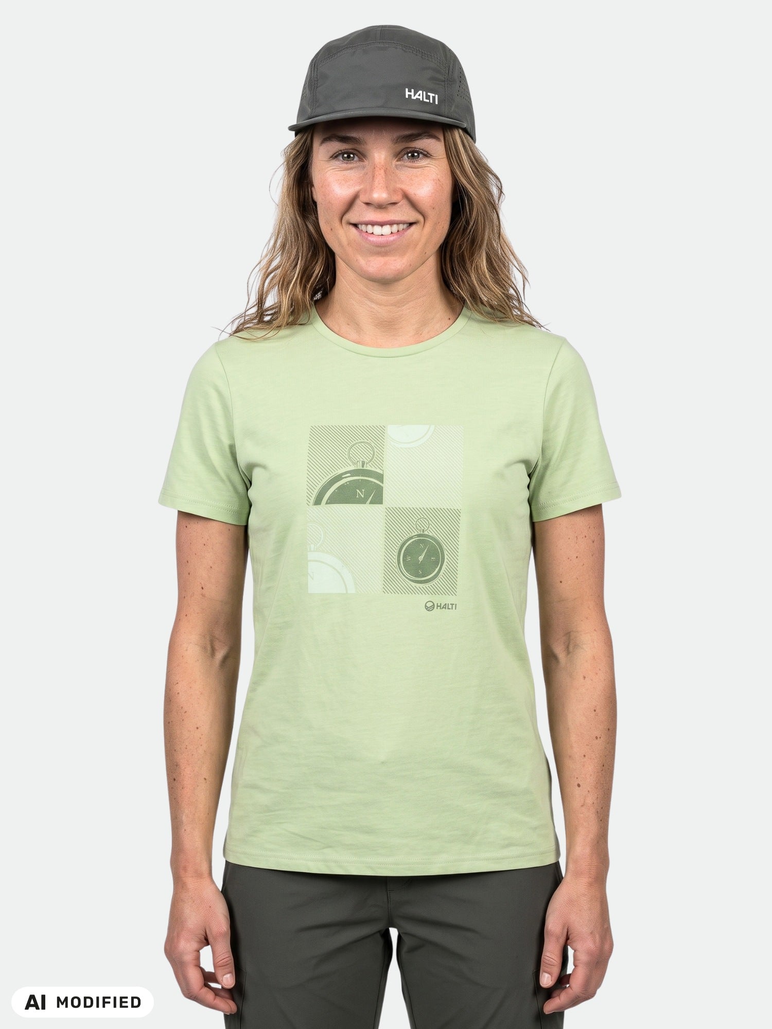 Woman wearing Halti's Matka T-shirt, a green organic cotton top with compass graphic, dark pants, and gray cap.