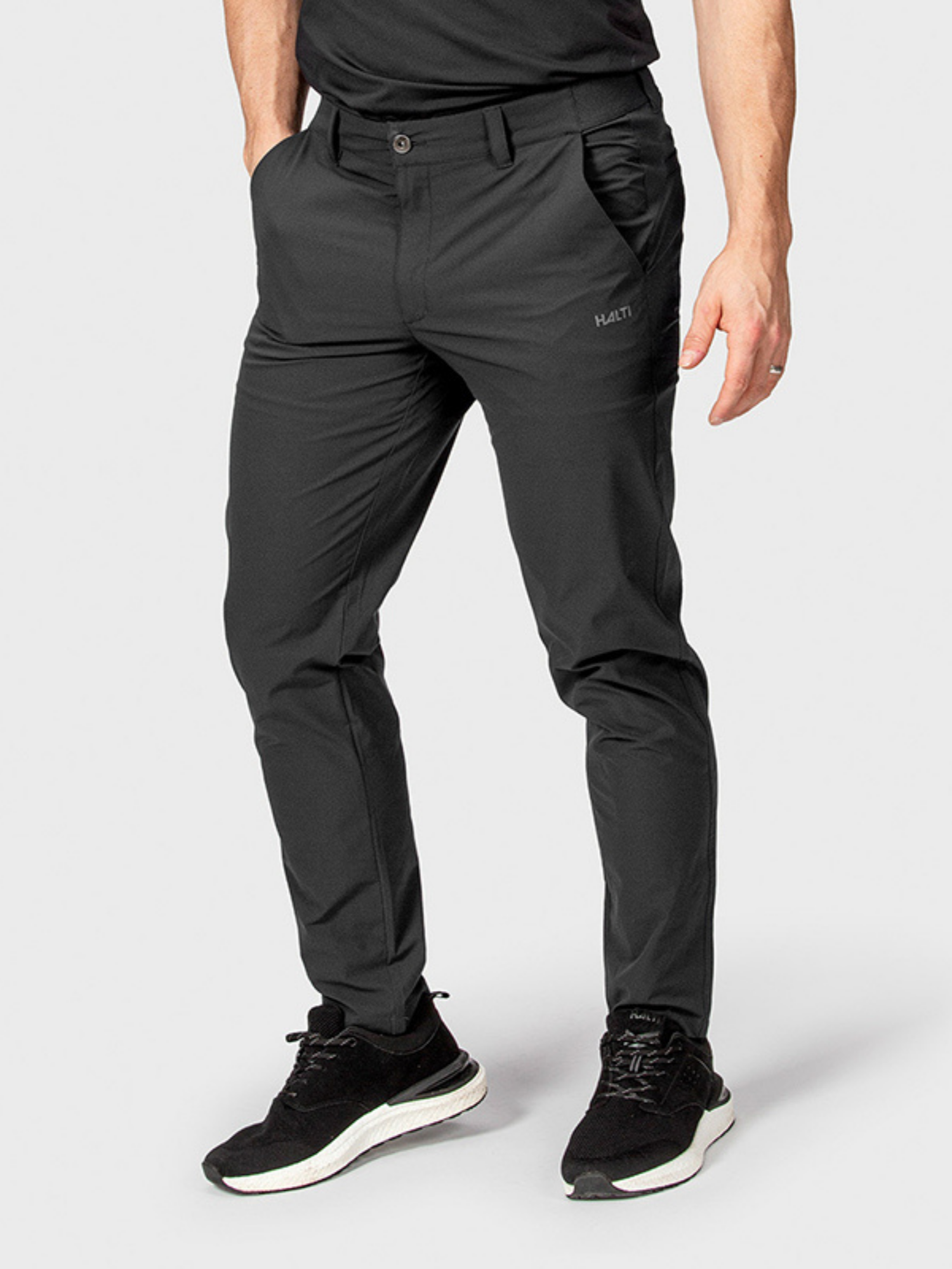 A person in Halti Drive Mens X-stretch Chinos and black sneakers stands hands-in-pockets against a light gray background.