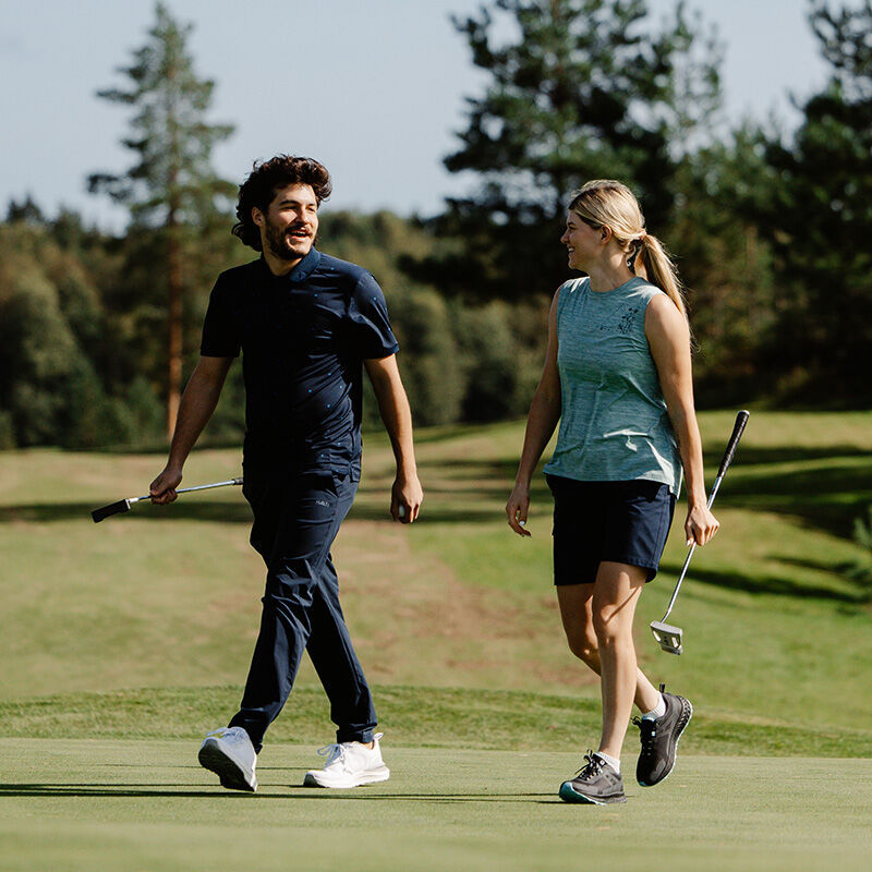 A man and woman in Halti Drive X-stretch Chinos walk on a golf course, each holding a putter among trees and grass.