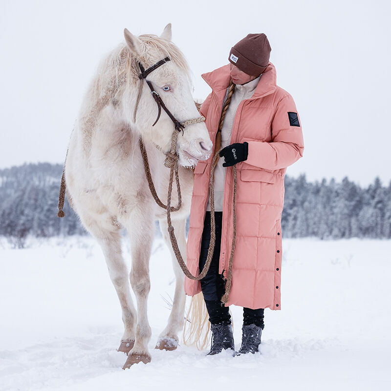 Woman in a Halti Penger Winter Jacket stands in snow, holding horses reins and looking at it.