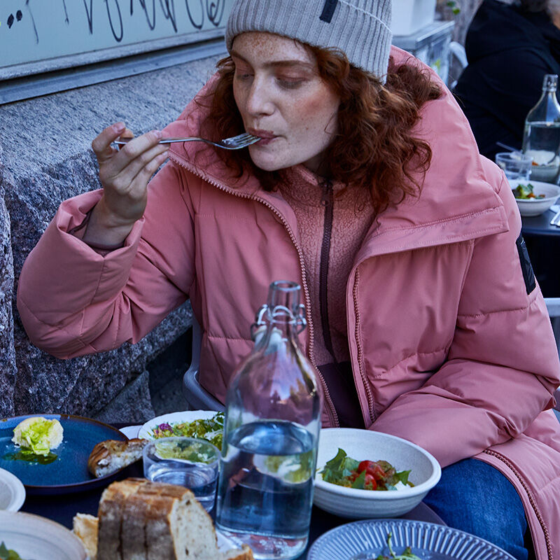 Wearing a pink Halti Penger Winter Jacket Womens and gray beanie, she eats salad, bread, and water outdoors.
