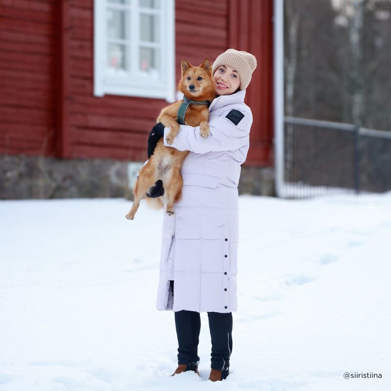 Wearing the Halti Penger Winter Jacket, a woman stands on snow holding a small dog in front of a red house.