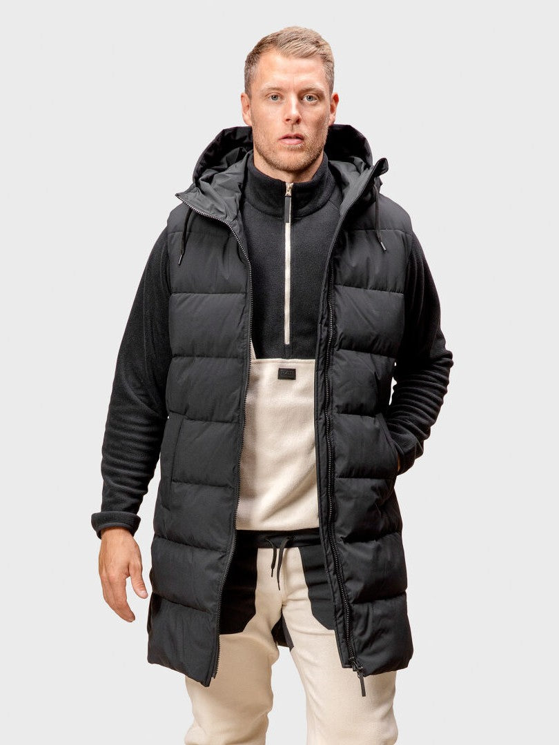 A man wears the Halti Penger Puffer Vest over a zip-up fleece and beige pants, standing facing forward.