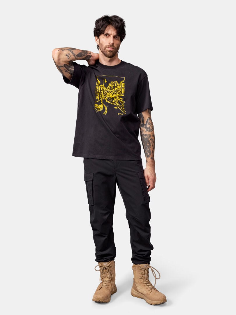Man standing with one hand behind his neck, wearing the Halti Skaltsi T-shirt Unisex, black cargo pants, and tan boots.