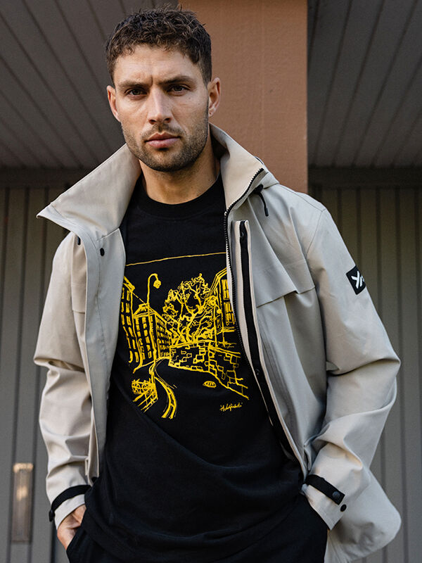 A man in a light jacket and Halti Skaltsi T-shirt Unisex stands outside by vertical wooden panels.
