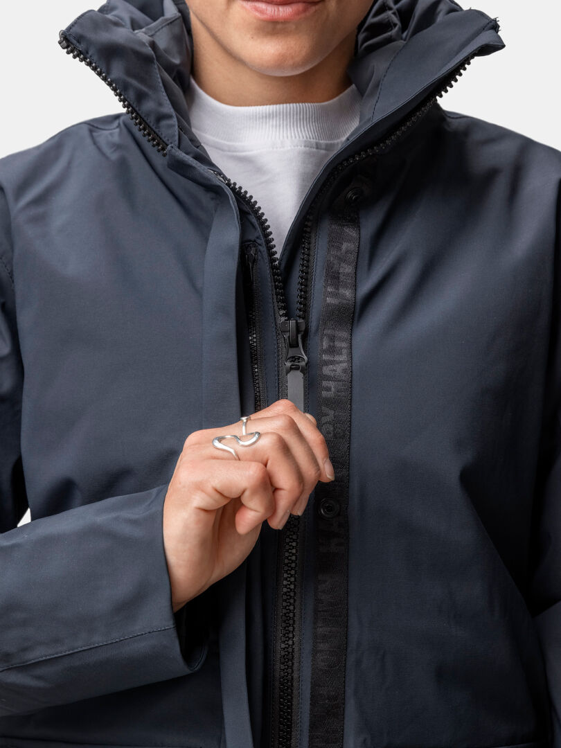 A woman zips up a Halti Synkas 3in1 DrymaxX Parka, wearing a white shirt and silver ring, shown shoulders down.