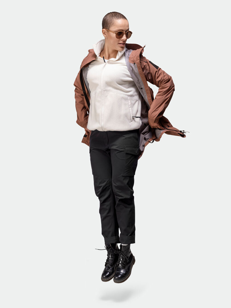 A woman wearing Halti Roams Cargo Pants jumps, styled with sunglasses, a brown jacket, and a white hoodie.