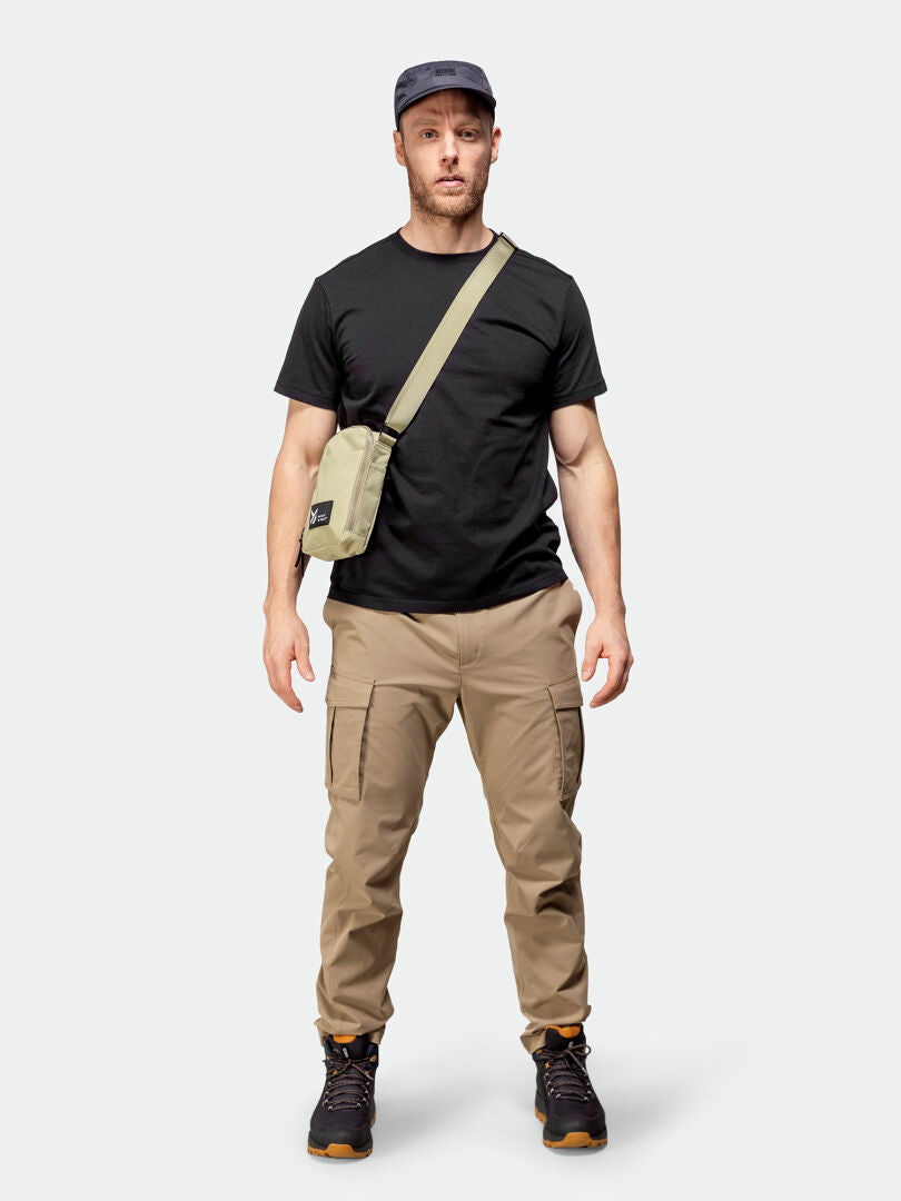 Man in Halti Roams Cargo Pants, black T-shirt, sneakers, cap, and crossbody bag against a plain background.