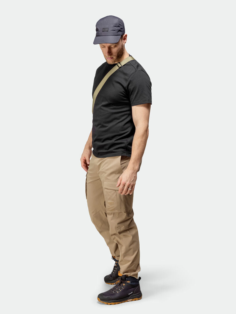 A man in Halti Roams Cargo Pants stands looking down, wearing a navy cap, black tee, and crossbody bag.