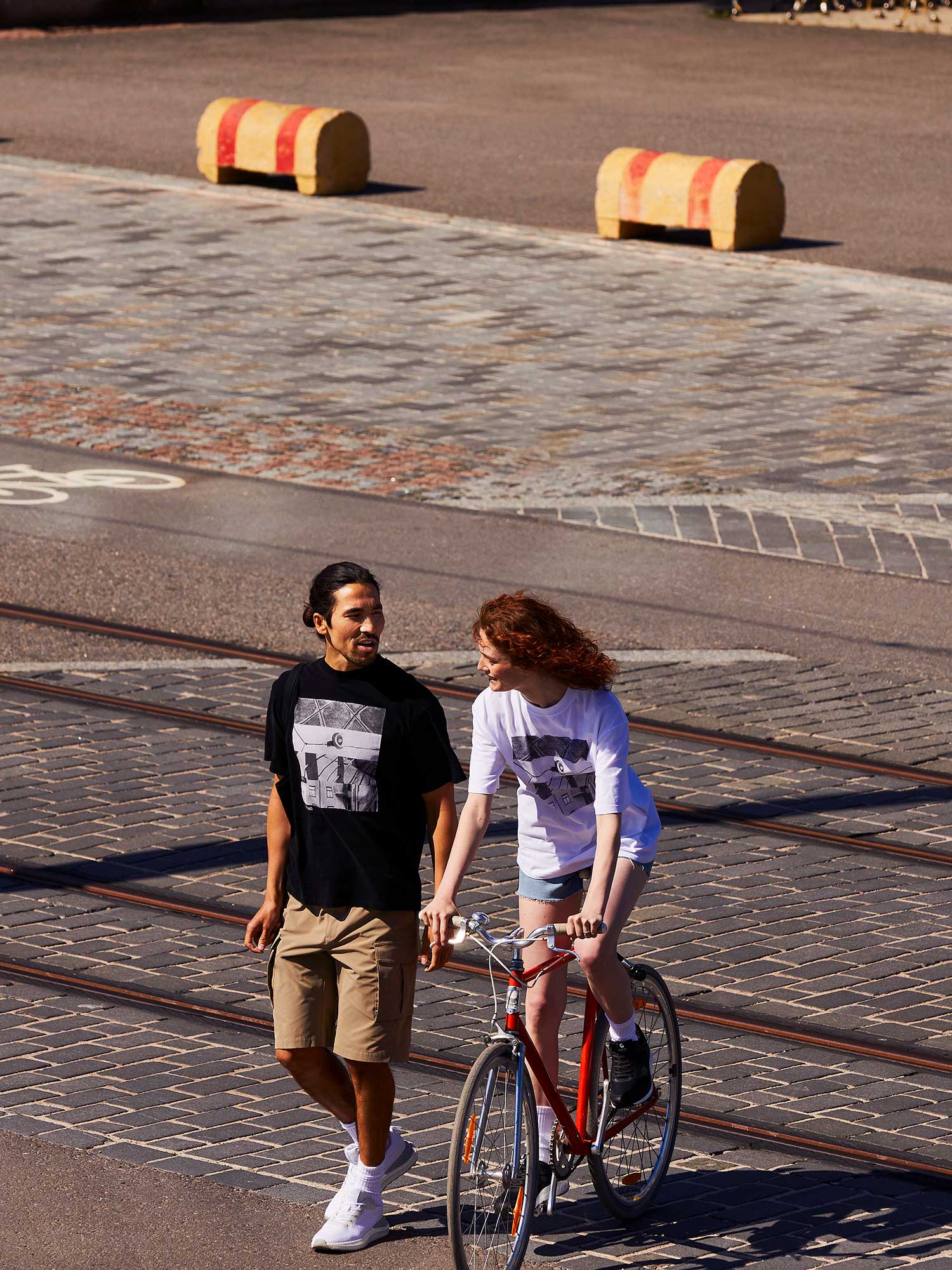 A man in Halti Roams Shorts walks beside a woman on a bike, both wearing graphic shirts on a cobblestone street.