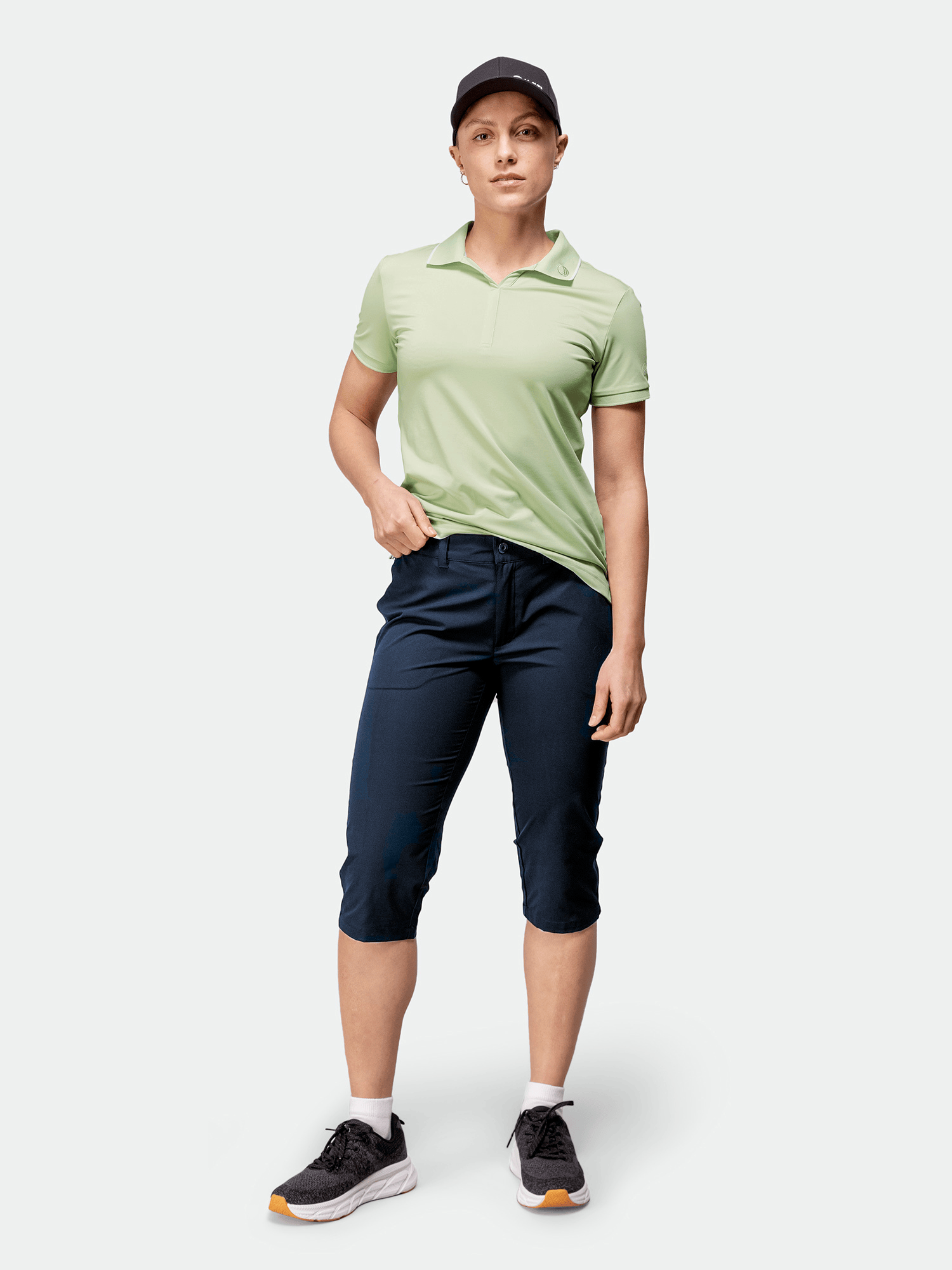 Woman in Halti Drive X-stretch Capri Pants, light green polo, black cap, and sneakers stands against a plain background.