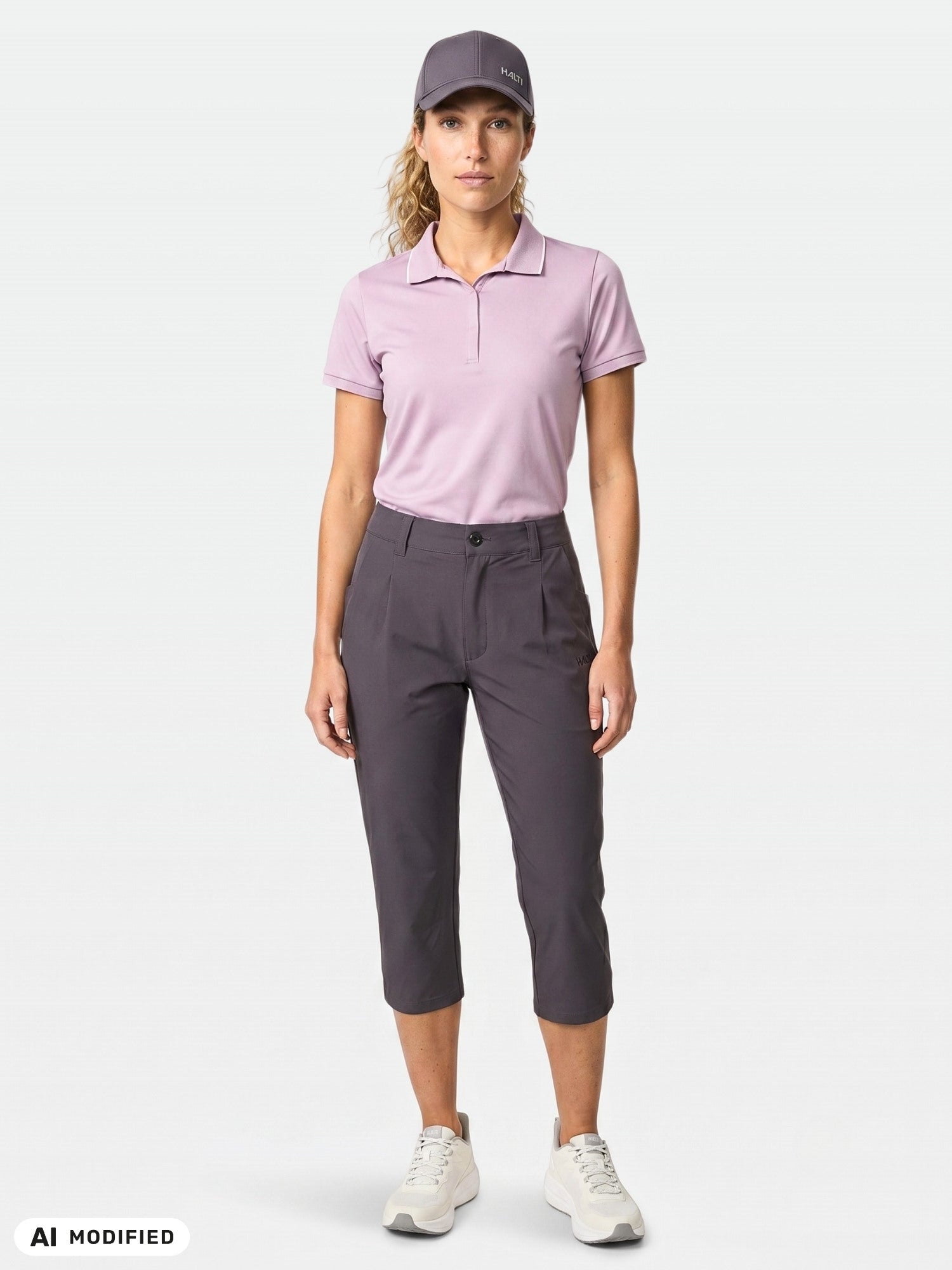 A woman faces forward wearing a lavender polo, purple cap, Halti Drive X-stretch Capri Pants, and white sneakers.