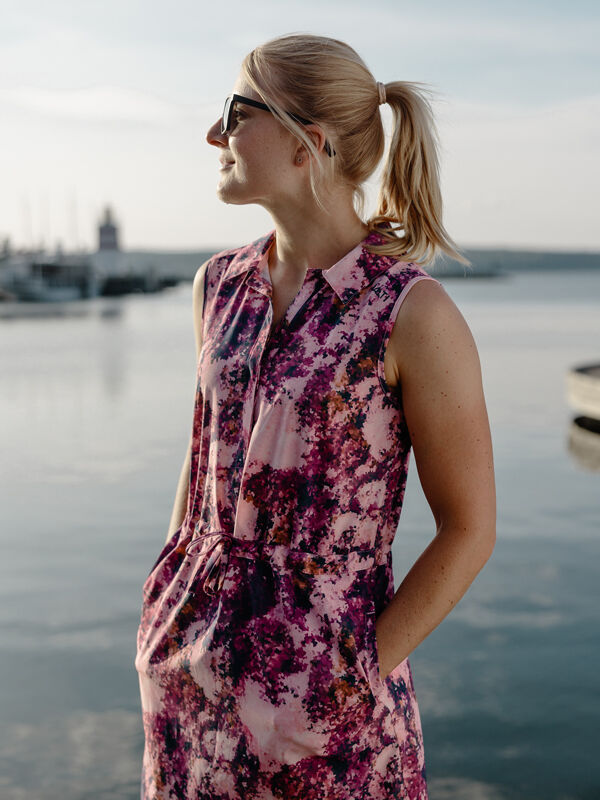 Blonde woman in ponytail wears Halti Chipper X-stretch Dress and sunglasses, standing by the water.