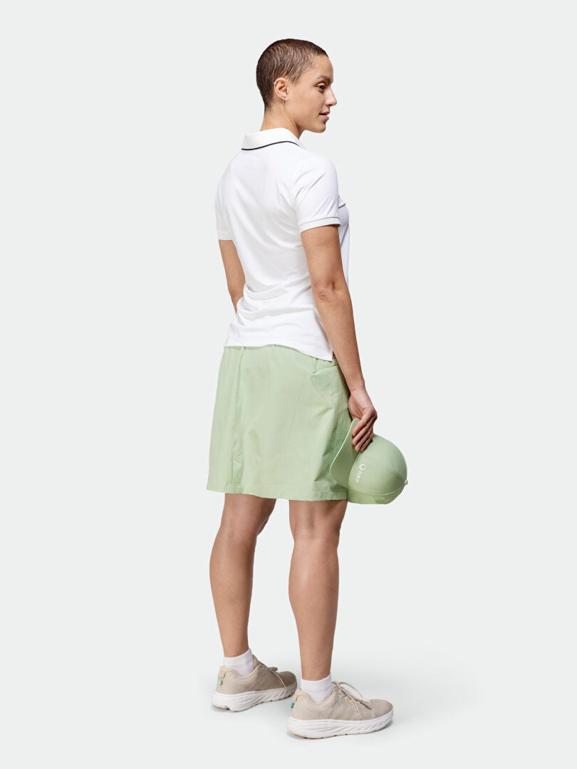 Woman facing backward in Halti Drive X-stretch Skort, white polo, beige sneakers, holding a light green cap.