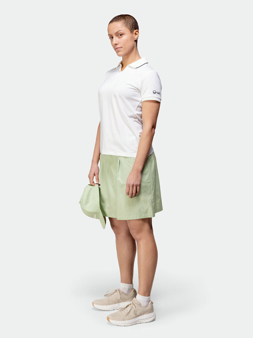Woman in Halti Drive X-stretch Skort light green, white polo, beige shoes, holding matching cap—outdoor ready.