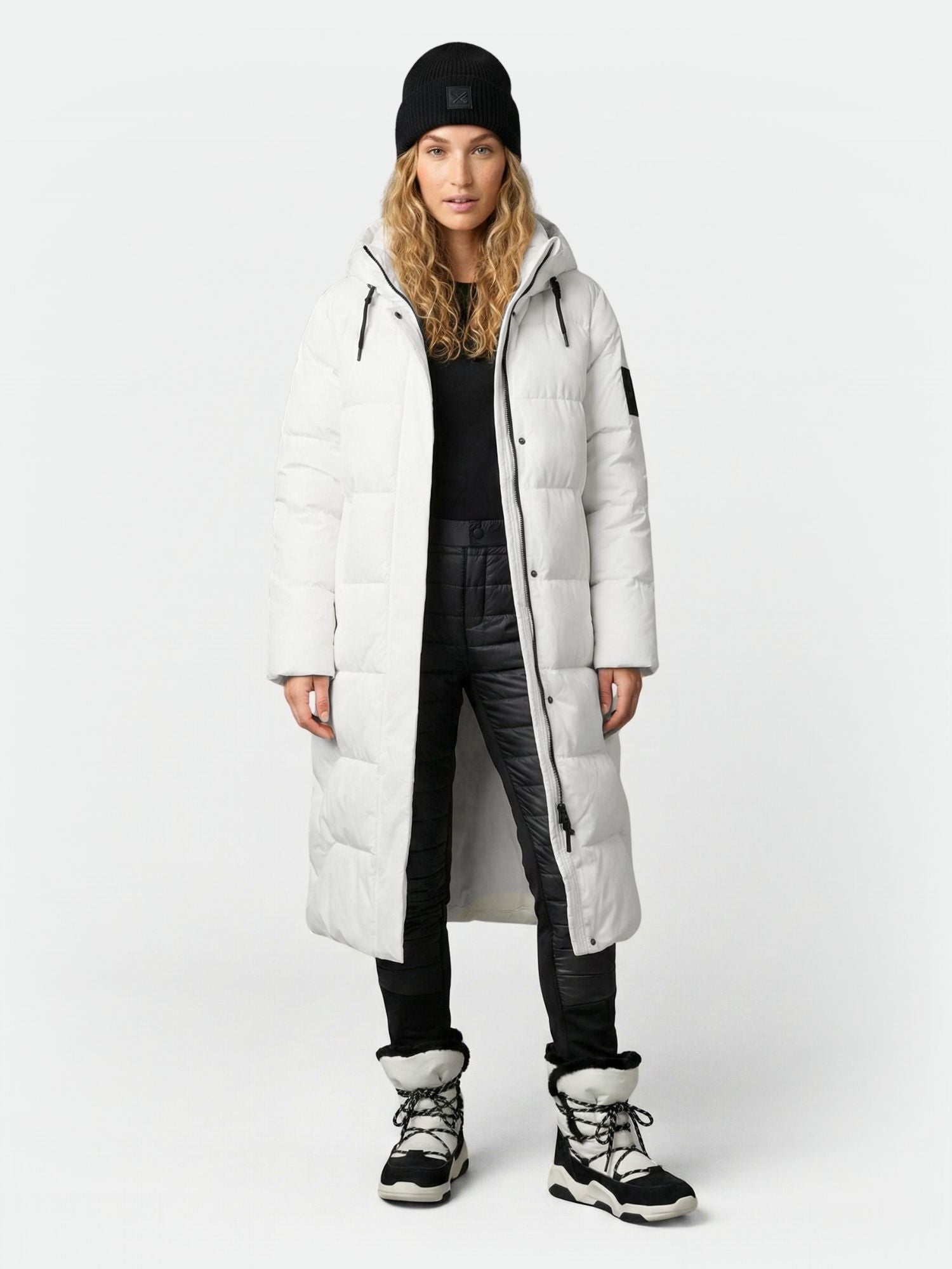 A woman faces forward wearing the Halti Brahe Puffer Long Winter Jacket in white, black pants, boots, and a beanie.