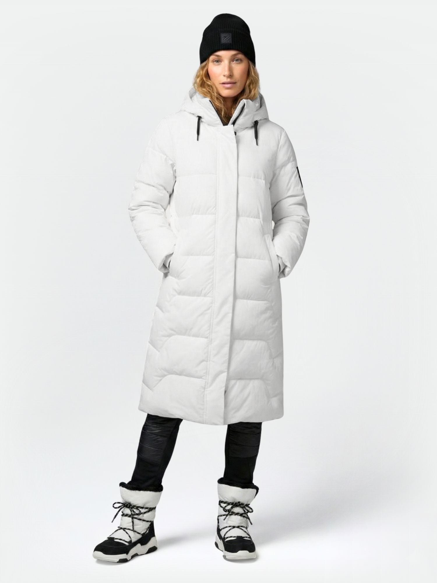 A person wears the Halti Brahe Puffer Long Winter Jacket for women, with a black beanie, black pants, and white boots.