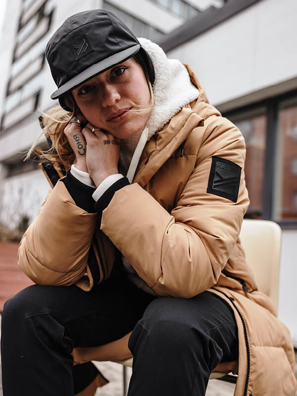 Woman in Halti Brahe Puffer Long Winter Jacket and black cap sits outdoors, leaning forward with hand on chin.