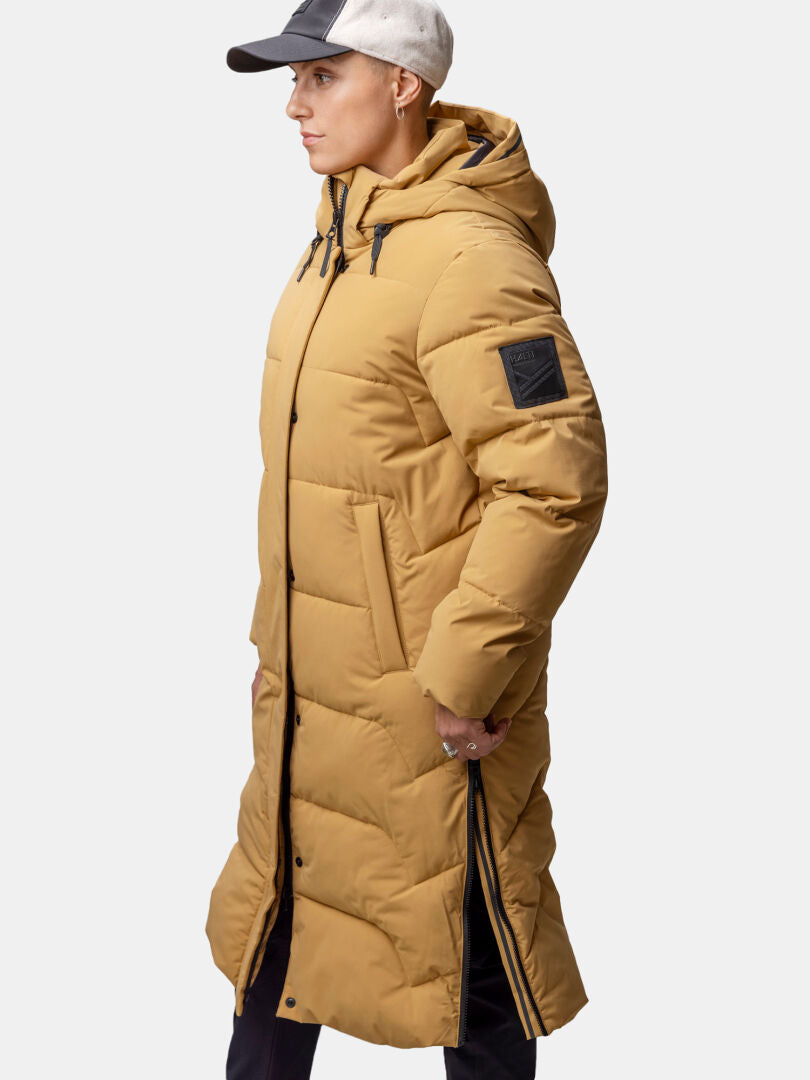 Woman wearing Halti Brahe Puffer Long Winter Jacket with hood, side zipper, sleeve patch, and cap, facing left.