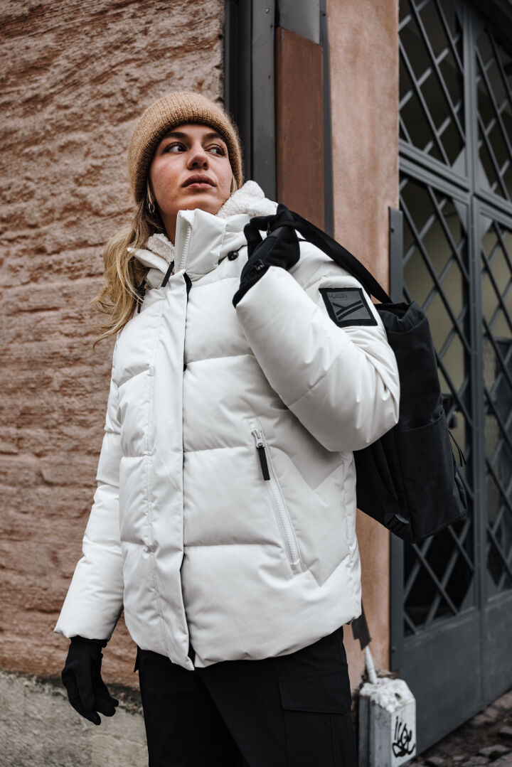 A person in the Halti Brahe Puffer Winter Jacket for women, black gloves, and beige beanie holds a backpack outdoors.