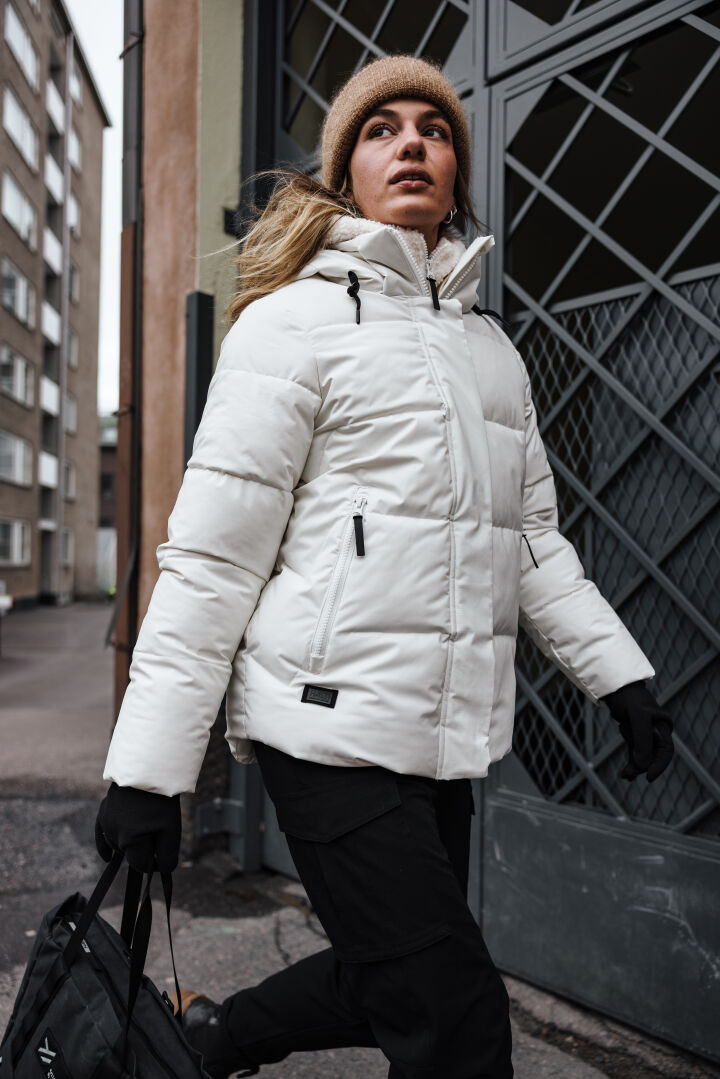 A person in a Halti Brahe Puffer Winter Jacket for women and tan beanie walks outside holding a black bag.