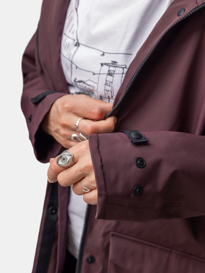 A person zipping up a maroon Halti Tokoi II DX Parka Jacket Womens, wearing silver rings and a white graphic tee.