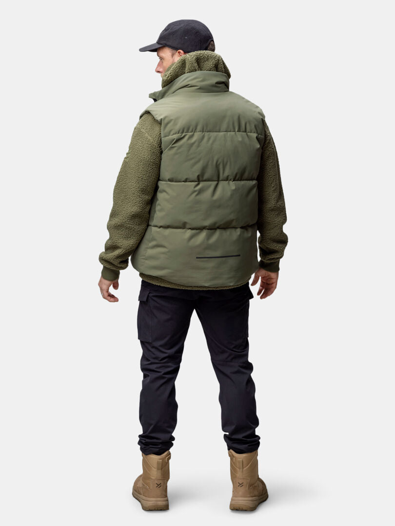 A man in a Halti Penger Classic Puffer Vest, olive fleece, black pants, tan boots, and a black cap faces away.
