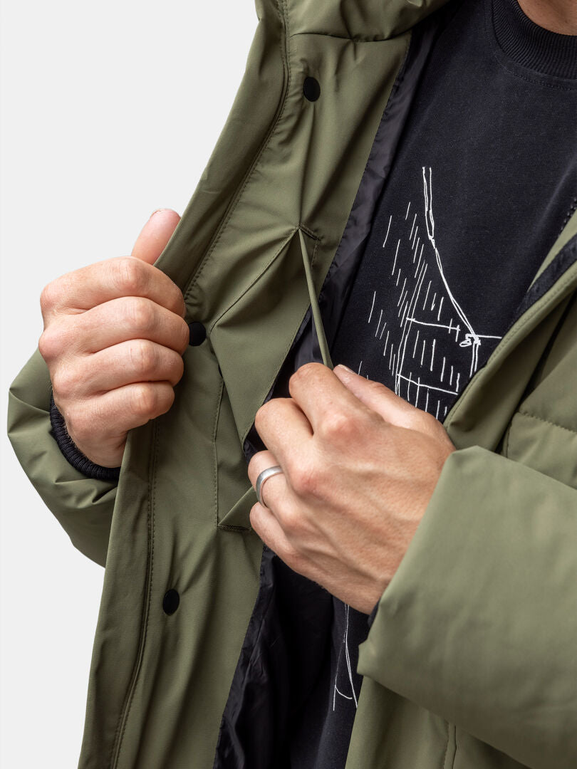 Someone adjusts the drawstring inside the Halti Brahe Puffer Winter Jacket Men's, worn over a black shirt with white lines.