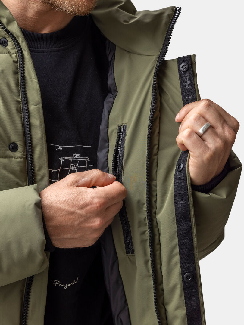 A man unzips the inner pocket of a Halti Brahe Puffer Winter Jacket, showing a black shirt beneath.