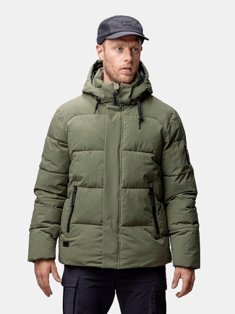 A man wears a Halti Brahe Puffer Winter Jacket Men's in olive green with a hood and black zipper, plus a black cap.
