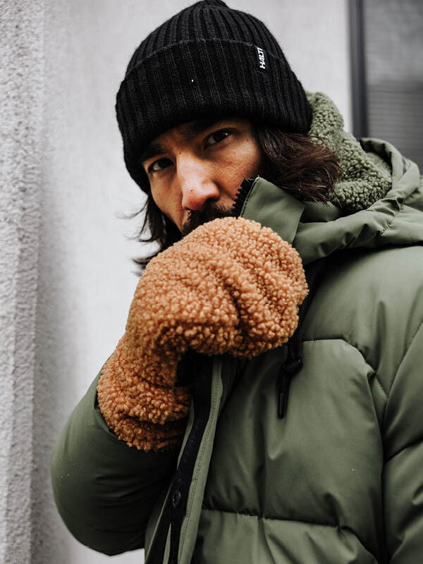 A man wears the Halti Brahe Puffer Winter Jacket, a black beanie, and brown mittens, standing outside near a wall.