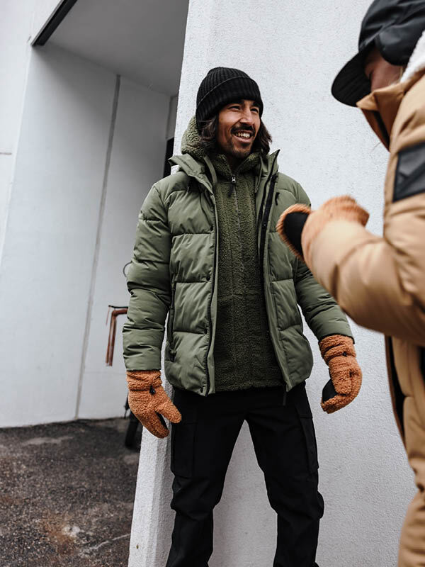 A man in a Halti Brahe Puffer Winter Jacket, olive hoodie, and black beanie smiles at someone outside.