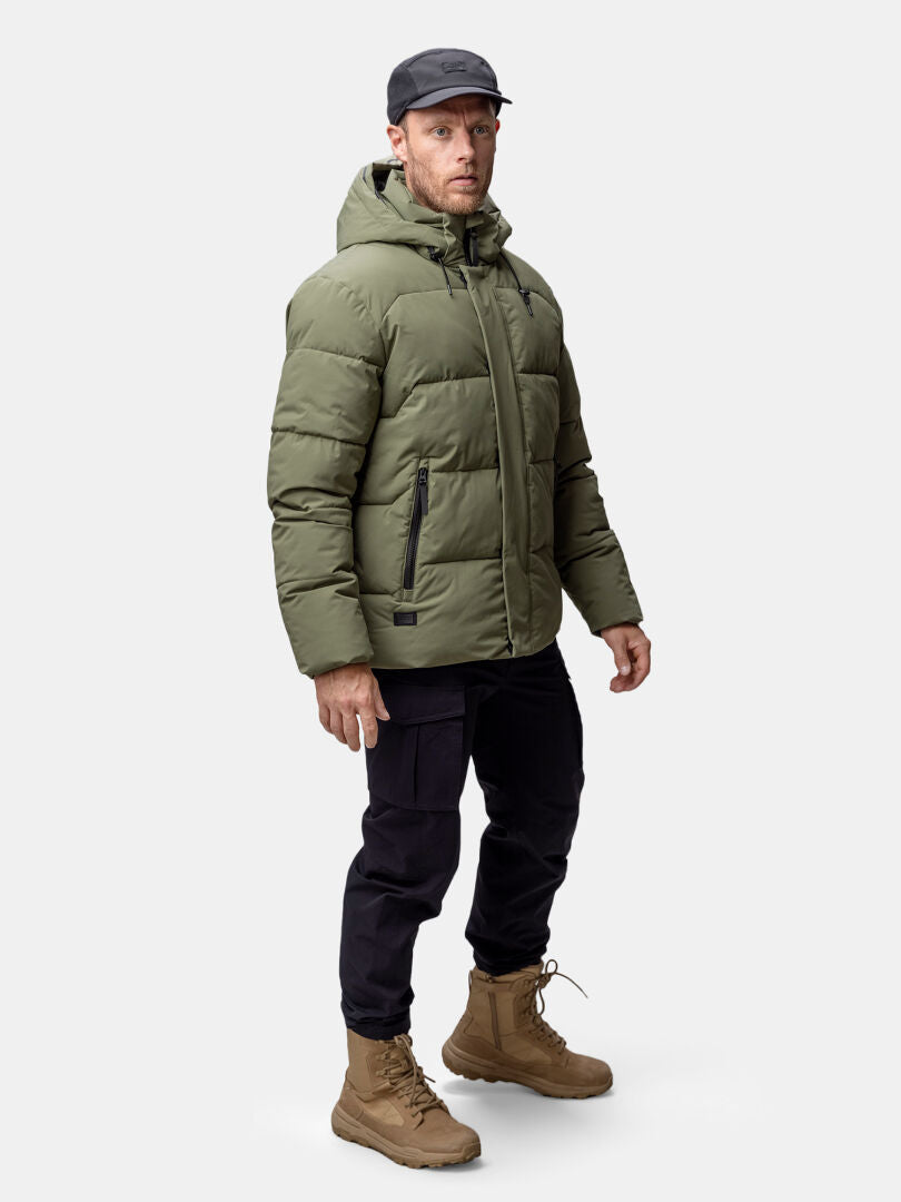 A man in a Halti Brahe Puffer Winter Jacket, black pants, tan boots, and a black cap stands against a plain background.
