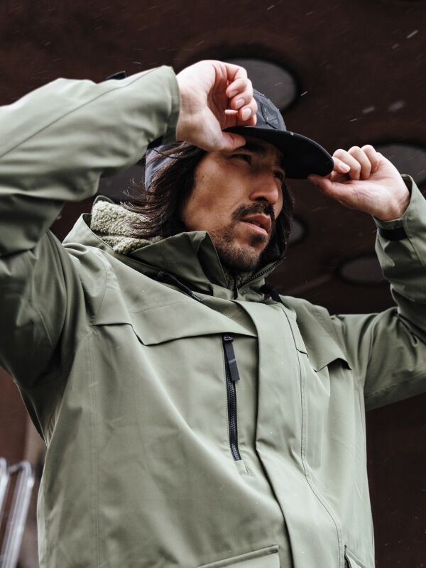 Man in a Halti Tokoi II DX Parka Jacket adjusts a black cap outdoors, looking up with a neutral expression.