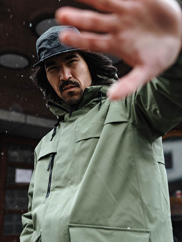 Man wears Halti Tokoi II DX Parka Jacket and black hat, standing outside with serious look, hand raised to camera.